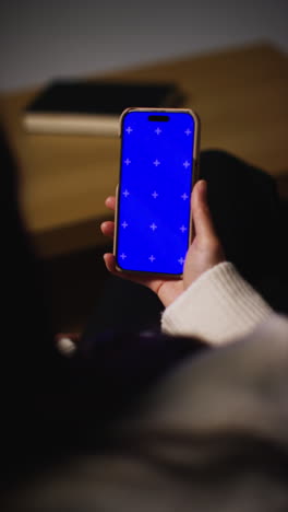 Vertical-Video-Over-The-Shoulder-Shot-Of-Woman-At-Home-Sitting-On-Sofa-With-Blue-Screen-Mobile-Phone-Scrolling-Through-Internet-Or-Social-Media